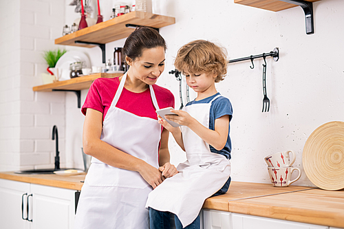Curly blond boy in apron showing his mother recipe in smartphone while both discussing it in the kitchen