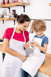 Cute little boy and his mother in aprons discussing ingredients of recipe in smartphone held by the child