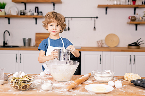 Cute little boy sifting flour over bowl while going to make dough for homemade pastry in the kitchen