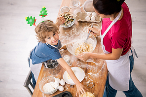 Adorable boy looking at you and stretching arm to dough while bending over wooden table in front of his mom cooking