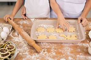 Hands of little boy with rolling-pin putting raw cookie in tray while helping mom with making cookies