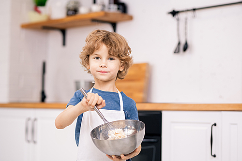 cute blond boy using whisker while mixing flour and eggs in steel bowl in front of camera in kitchen