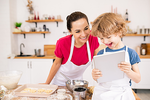 Adorable joyful child with touchpad showing his mom video recipe of something really yummy while choosing what to cook