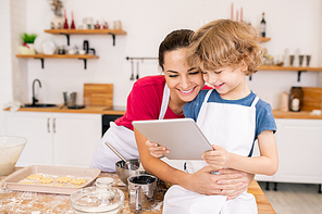 Adorable boy with touchpad and his happy mom discussing video recipe while choosing what to cook for dinner