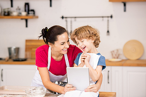 Joyful boy and his mother in aprons laughing at video in touchpad while sitting in the kitchen and choosing what to cook