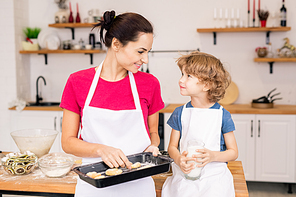 Happy young woman holding tray with baked cookies while looking at her little son and talking to him in the kitchen