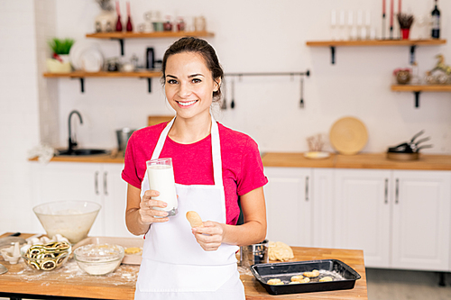 Pretty young brunette female in apron holding glass of milk and baked homemade cookie while enjoying lunch