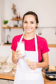 Happy young brunette female with toothy smile having glass of milk with baked homemade cookie in front of camera