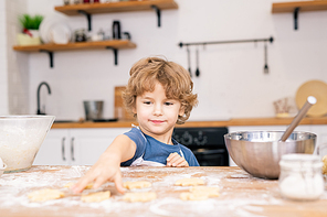 Cute little boy in apron stretching hand to one of raw cookies on table while helping his mom with cooking