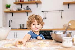 Smiling little boy looking at you while going to take one of raw cookies on table while helping his mom