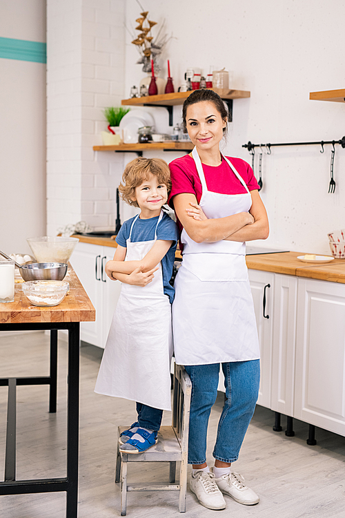 Happy adorable child and his mother in white aprons standing close to one another in the kitchen