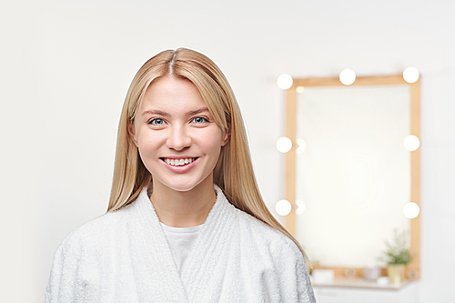 Pretty young blonde female with toothy smile standing in front of camera on background of mirror