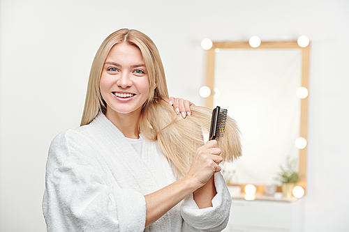 Happy and healthy young woman with toothy smile brushing her long blond hair in front of camera