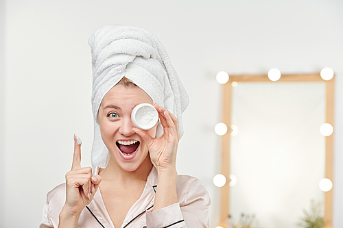 Ecstatic girl with white towel on head holding jar of facial moisturizer by left eye while keeping forefinger raised