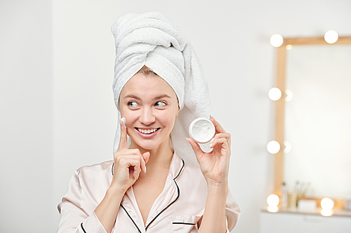 Fresh young beautiful woman with white towel on head applying her facial hydrating cream on cheek after cleaning face