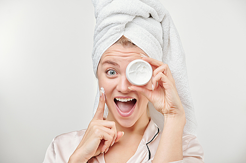 Excited girl with towel on head holding jar of facial hydrating cream by left eye while applying the product on cheek