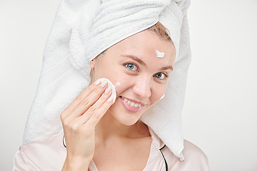 Pretty young healthy woman using cotton pad for applying hydrating cream on face while standing in front of camera