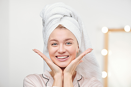 Pretty cheerful girl with towel on head enjoying her healthy skin while keeping open palms by face after morning hygiene