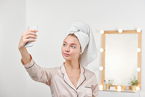 Fresh young female in silk pajamas making selfie after shower while standing on background of mirror with lamps