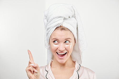 Happy beautiful woman with under-eye patches on face and towel on head pointing upwards in isolation