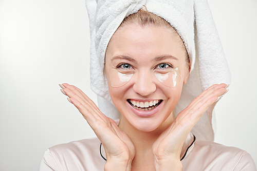 Young beautiful woman with under-eye patches on face and towel on head looking at you with toothy smile