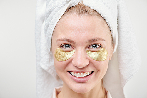 Healthy cheerful girl with towel on head and revitalising golden under-eye patches standing in front of camera