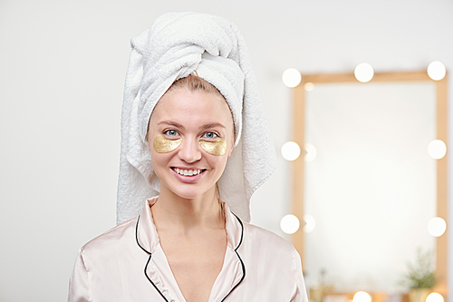 Pretty young smiling female with towel on head having under-eye mask in bathroom after shower