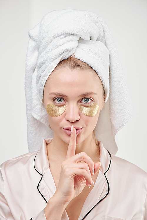 Fresh and clean young woman with towel on head and golden under-eye patches keeping forefinger by her mouth