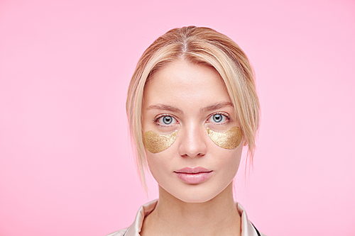 Serious young blond woman with golden revitalising under-eye patches standing against pink background