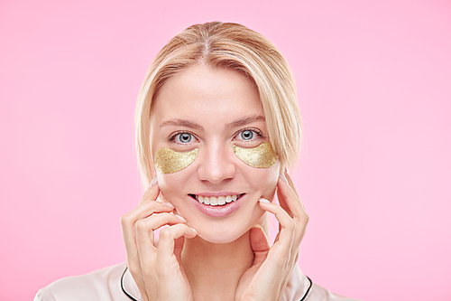 Smiling blond girl with golden revitalising under-eye patches touching her face while standing against pink background