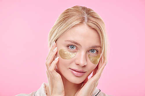 Young beautiful blond woman with golden revitalising under-eye patches keeping hands on her radiant face