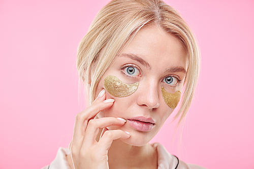 Attractive blond woman touching her face during skincare procedure for under-eye area against pink background