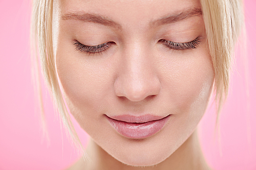 Face of young blond serene woman with natural makeup looking down in front of camera over pink background