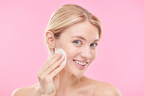 Pretty young smiling female with cotton pad applying toner or micellar water while cleansing her face against pink background