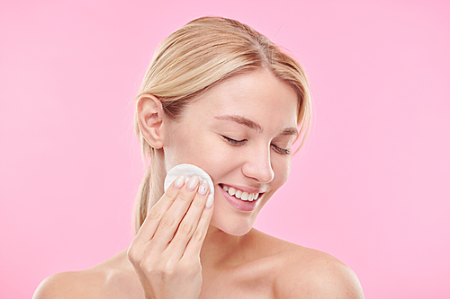 Pretty young smiling female with cotton pad enjoying procedure of cleaning face with toner or micellar water