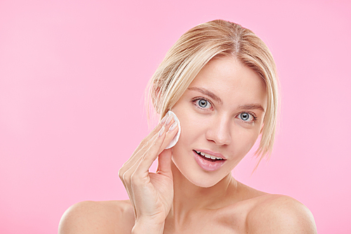 Beautiful blond girl using cotton pad while cleansing face and removing makeup over pink background