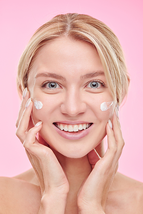 Young cheerful woman with toothy smile pampering cream on her face after cleaning it with toner