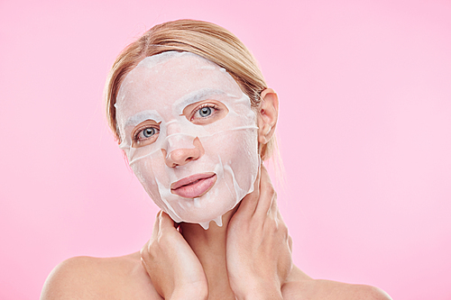 Blond girl with textile moisturizing mask on face looking at you while keeping hands on neck in isolation
