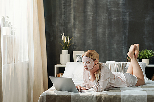 Young busy female in silk pajamas lying on bed in front of laptop while taking care of her face and working at home