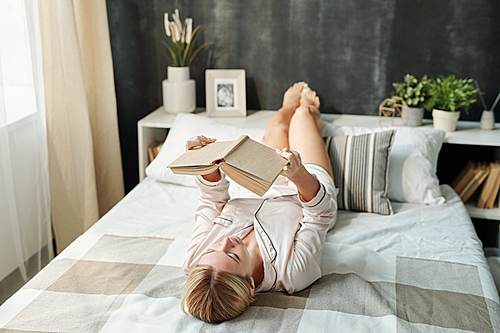 Blond girl in pajamas lying on bed and reading book while preparing for exam or for pleasure