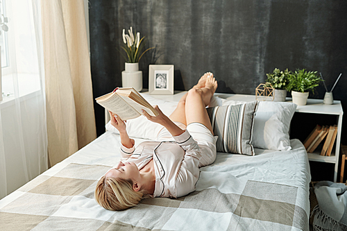 Pretty young woman in pajamas relaxing on bed at leisure and reading book while staying at home on weekend