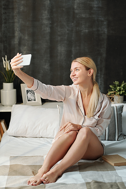 Joyful blond girl in silk pajamas looking at smartphone camera while making selfie on bed in the morning