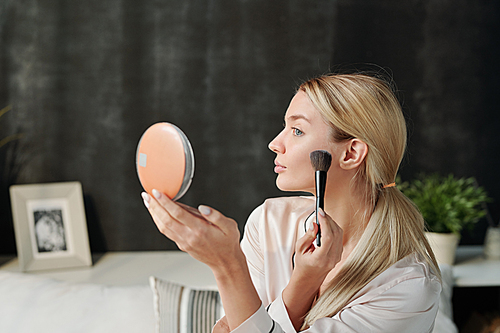 Pretty blond woman applying powder on her face with brush while looking in mirror during makeup process