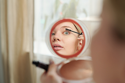 Reflection in mirror of young woman applying black mascara on eyelashes while doing makeup in the morning