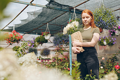 Pensive attractive young redhead woman in casual clothing holding flowers in kraft paper and choosing flowers in greenhouse