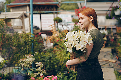 Smiling beautiful redhead girl holding lilies in paper bag and looking around, she growing flowers in greenhouse
