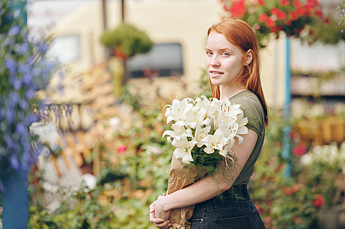 Portrait of content attractive redhead girl working with flowers at plant market, she holding white lilies in paper