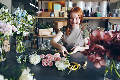 Cheerful beautiful redhead florist in apron standing at table with tender flowers and arranging bouquet in flower shop