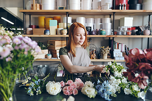 Content experienced young woman in apron standing at desk and choosing fresh flowers for bouquet in flower shop