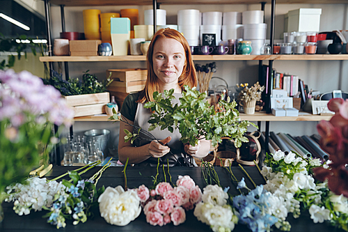 Portrait of smiling attractive redhead girl in apron standing at table with flowers in row and adding green branches to bouquet